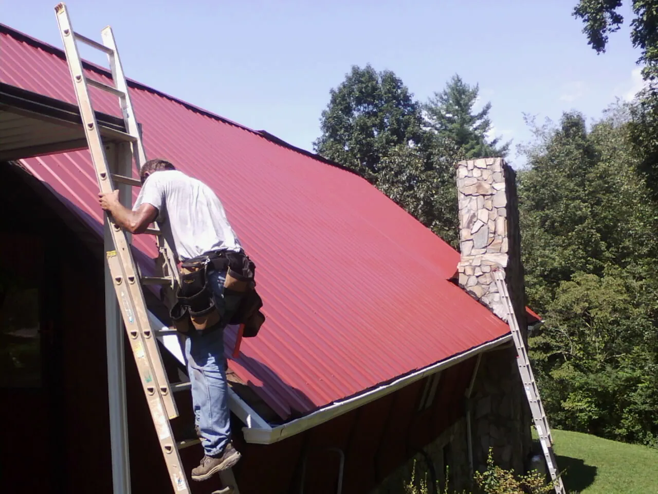 Roofer on a ladder beside a red metal roof and stone chimney — Soffit & Fascia Repair by Roofing Brothers in Edna